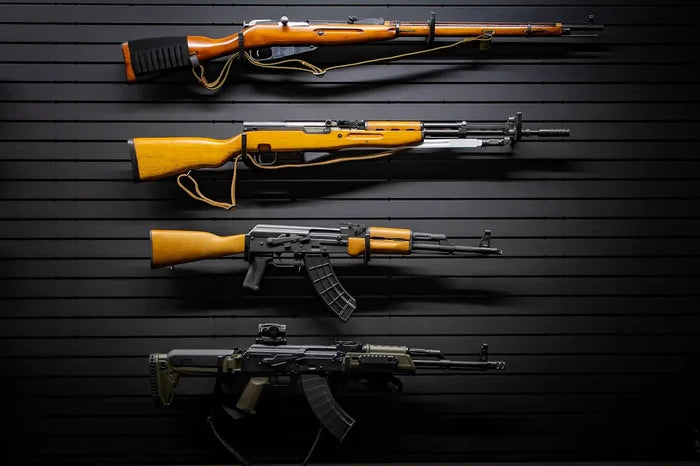 Four rifles displayed horizontally on a black Tactical Walls ModWall panel using multiple MWHRACKRS rack pairs: a Mosin-Nagant bolt-action rifle, an SKS rifle with wooden stock, a wooden-stock AK-47 with 30-round magazine, and a tactical AK variant with green furniture and red dot sight